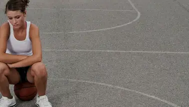 A young woman in a tank top sitting on a basketball in the middle of an outdoor court, looking down