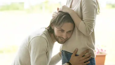 Homme accroupi qui pose l'oreille sur le ventre arrondi d'une femme enceinte debout, la main de celle-ci dans ses cheveux