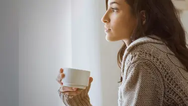 Femme brune en gilet tricoté, de profil devant une fenêtre claire, une tasse blanche entre les mains