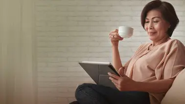 A middle-aged woman sitting cross-legged with a tablet on her lap and a raised cup, against a white brick wall