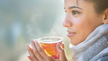 Close-up of a woman in a grey scarf holding a steaming cup of tea close to her face, outdoors