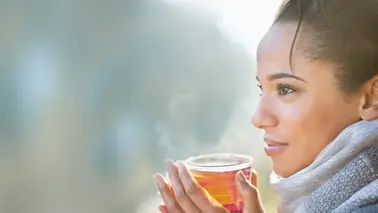 Jeune femme au foulard gris, tournée vers la lumière, qui tient à deux mains une tasse de thé fumante