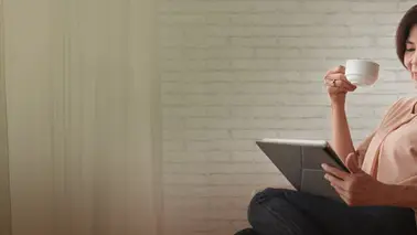 A middle-aged woman in a pink blouse reading on a tablet, cup raised, against a white brick wall