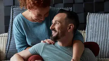 A middle-aged couple on a sofa: the red-haired woman laughs as she leans over the bearded man, who looks up at her