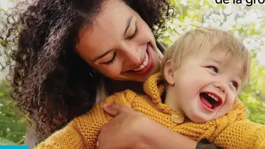 Une mère aux cheveux bouclés serre un bébé rieur en chandail moutarde, entourés d'arbres en fleurs