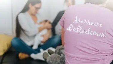 Personne de dos portant un chandail rose où on lit « Marraine d'allaitement »; à l'arrière-plan flou, une femme allaite un bébé